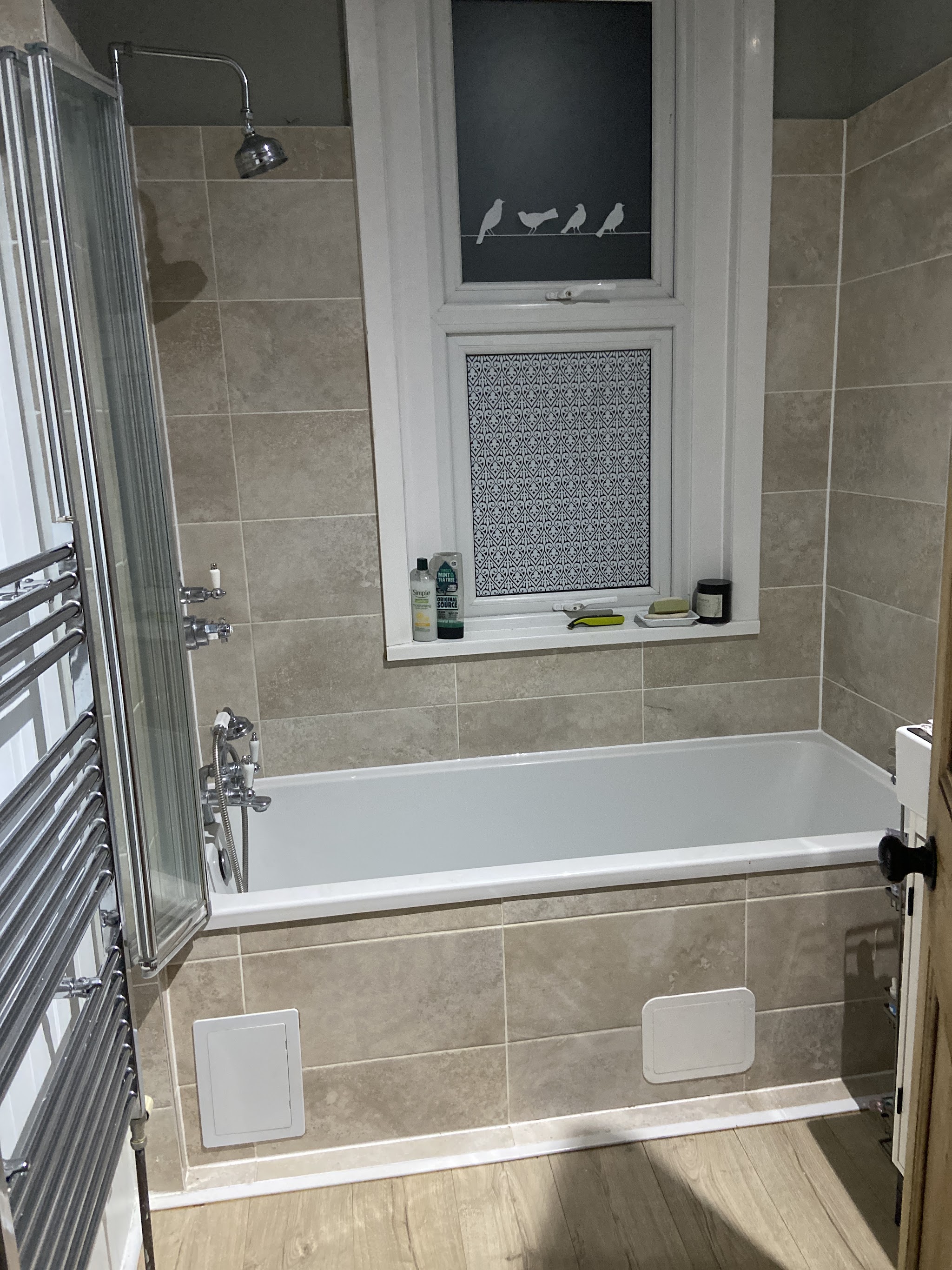 A finished bathroom with a white bath, glass shower screen, beige tiles and a chrome towel radiator