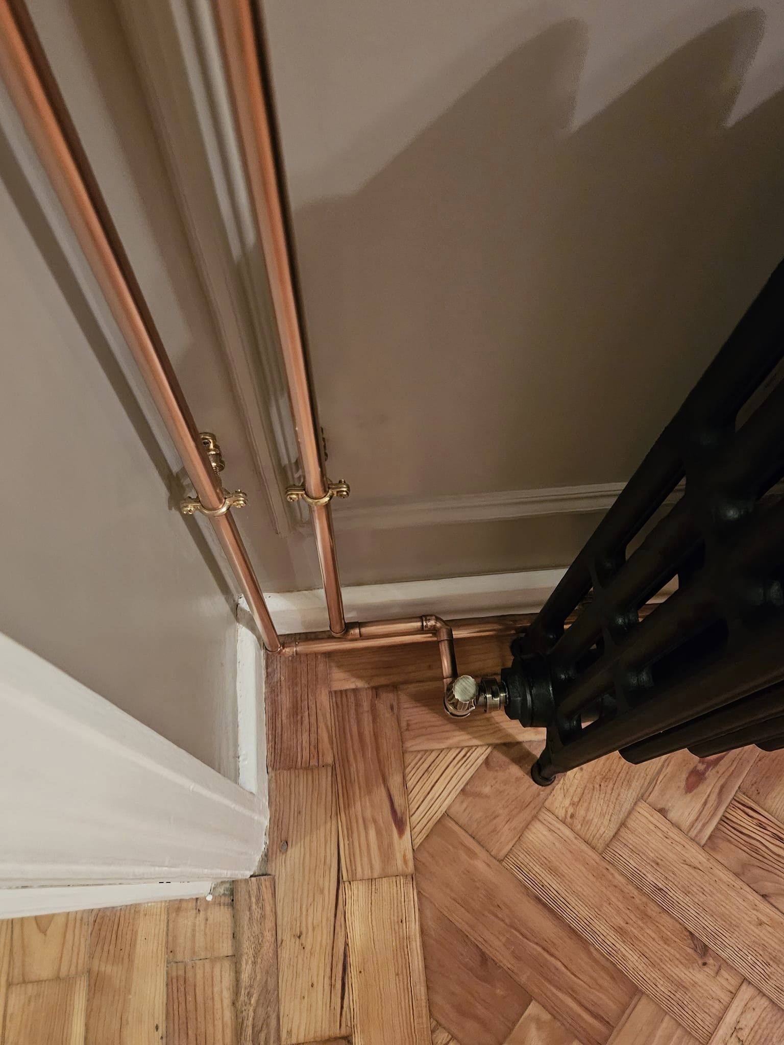 Copper pipework dropping down a wall to a black cast-iron column radiator on a herringbone floor